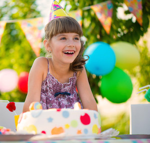 Little girl smiling at a birthday party, sitting in front of a cake and wearing a birthday hat with balloons in background